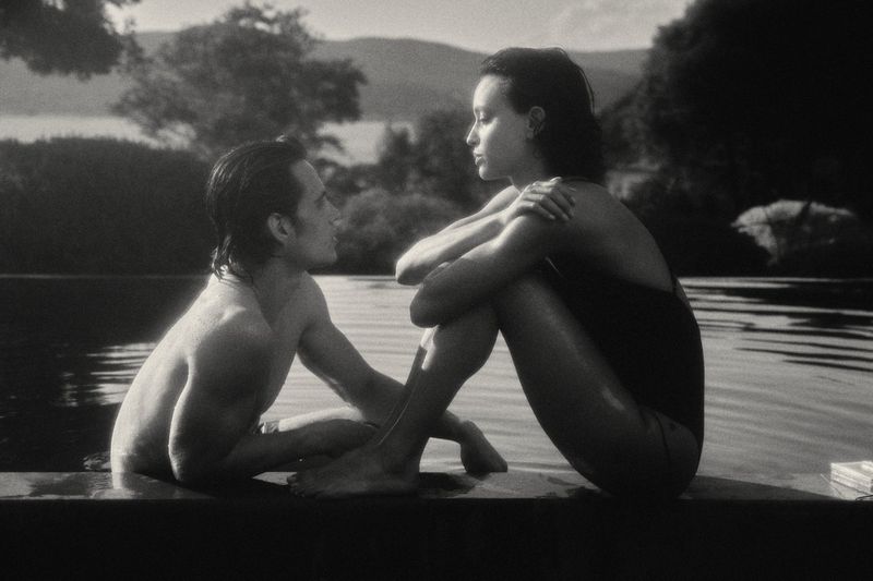 Monochrome image of a couple seated poolside at an infinity pool, gazing at each other with a mountain range in the background.