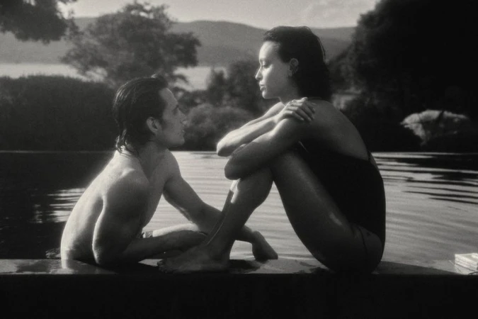 Monochrome image of a couple seated poolside at an infinity pool, gazing at each other with a mountain range in the background.