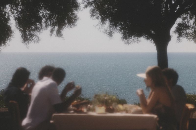 People at an outdoor table with a view of the ocean, under the shade of a tree. The focus is soft, creating a dreamy atmosphere.