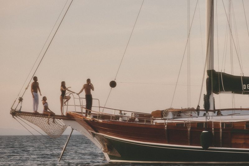 A group of four people on the bow of a wooden sailboat, with the vast ocean stretching behind them under a soft, warm sky.