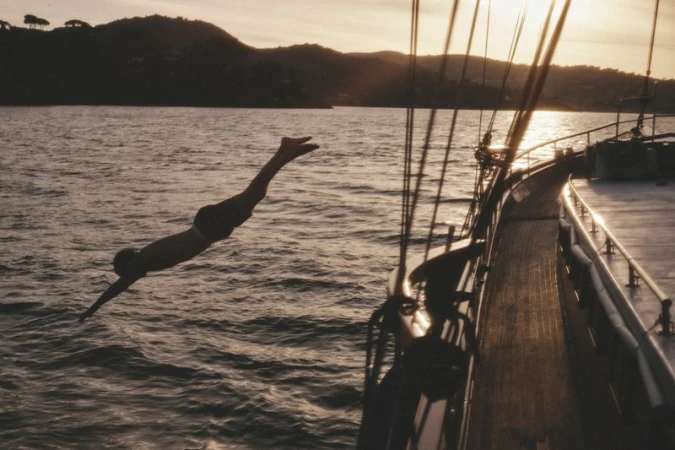 Person dives into rippled ocean water from the side of a sailboat with visible rigging; sunset and distant land, silhouetted against the sky.