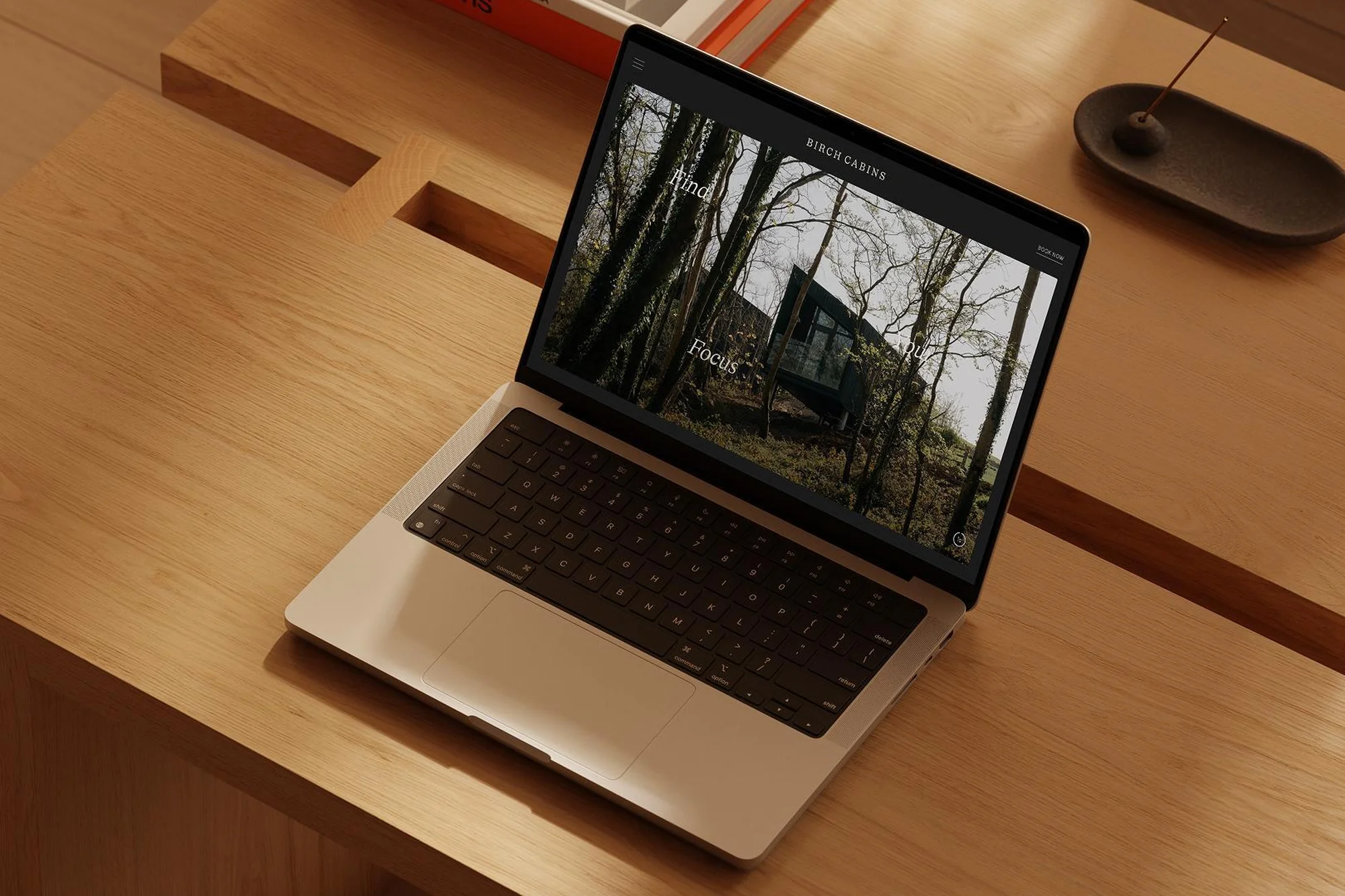 Laptop displays a webpage showcasing a cabin in the woods, resting on a light wood table next to a dark incense holder. Minimalist.