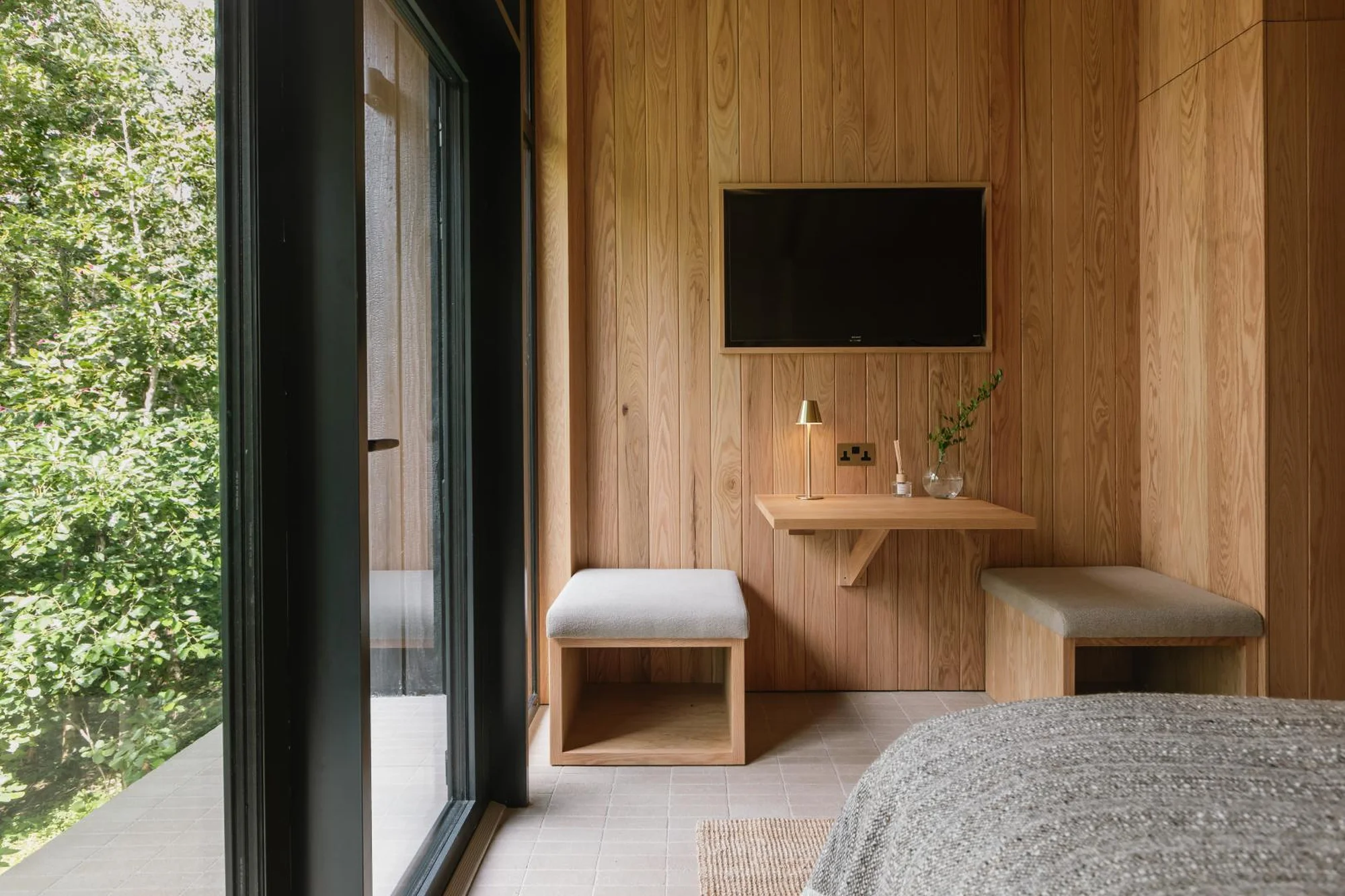 Contemporary bedroom interior with wood paneling, a TV, and a view of a green forest through a large window, bringing nature indoors.