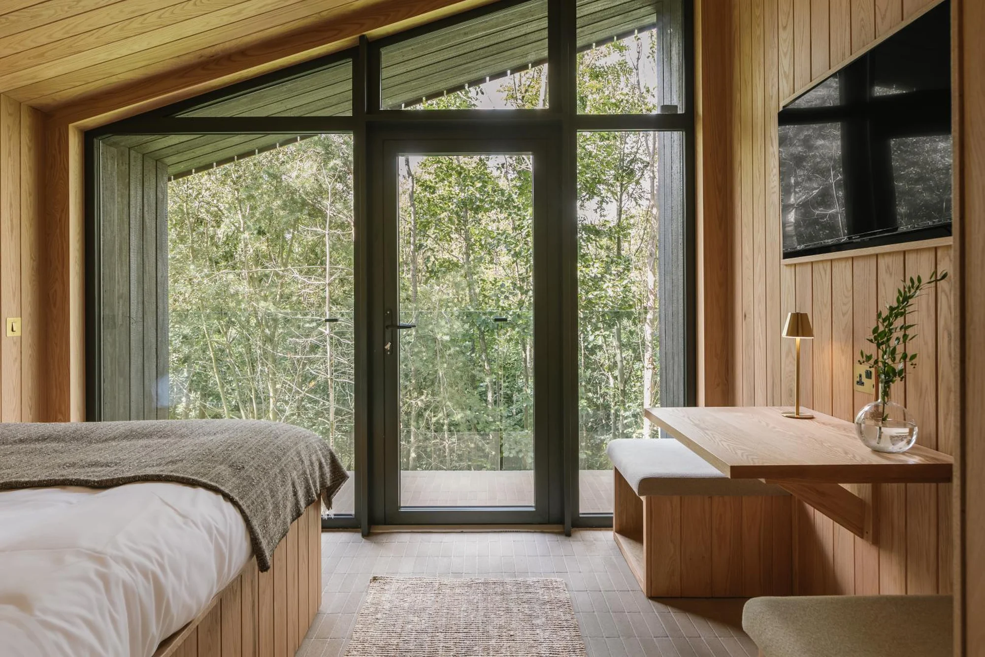Bedroom with wood paneled walls and ceiling, large windows overlooking trees, a bed, desk with lamp, and a television.
