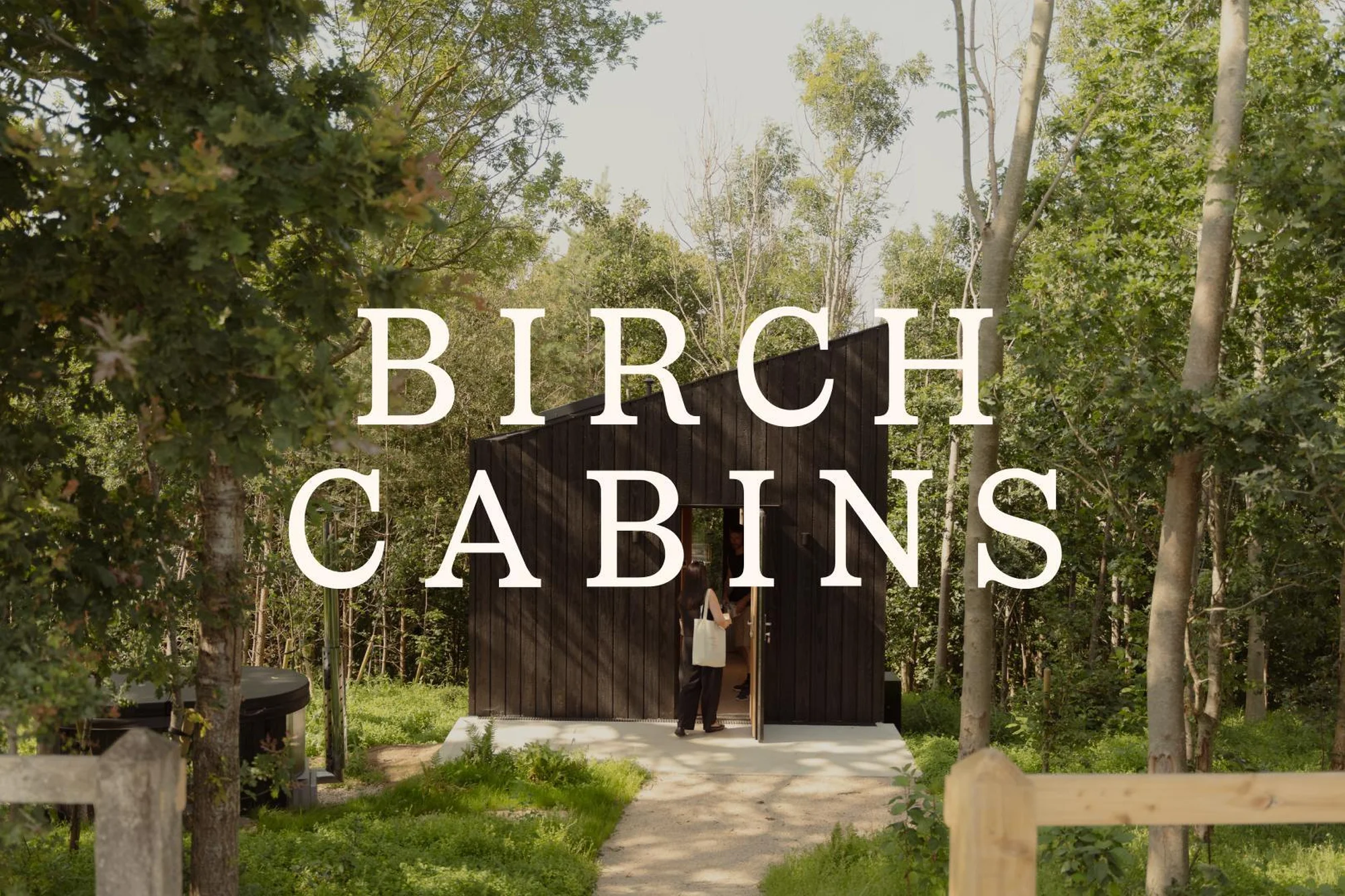 Exterior view of a modern cabin with a woman entering, framed by a forest of birch trees and a path leading to the entrance.