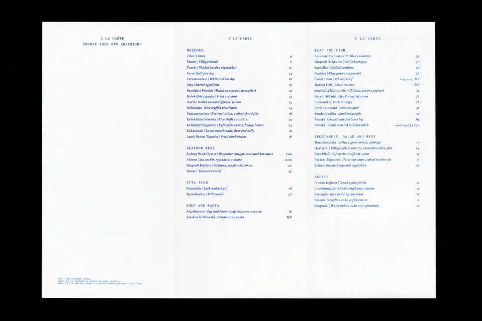 Bright studio shot of a tri-fold restaurant menu with Greek cuisine options including seafood, meat, vegetables, and desserts in blue ink.