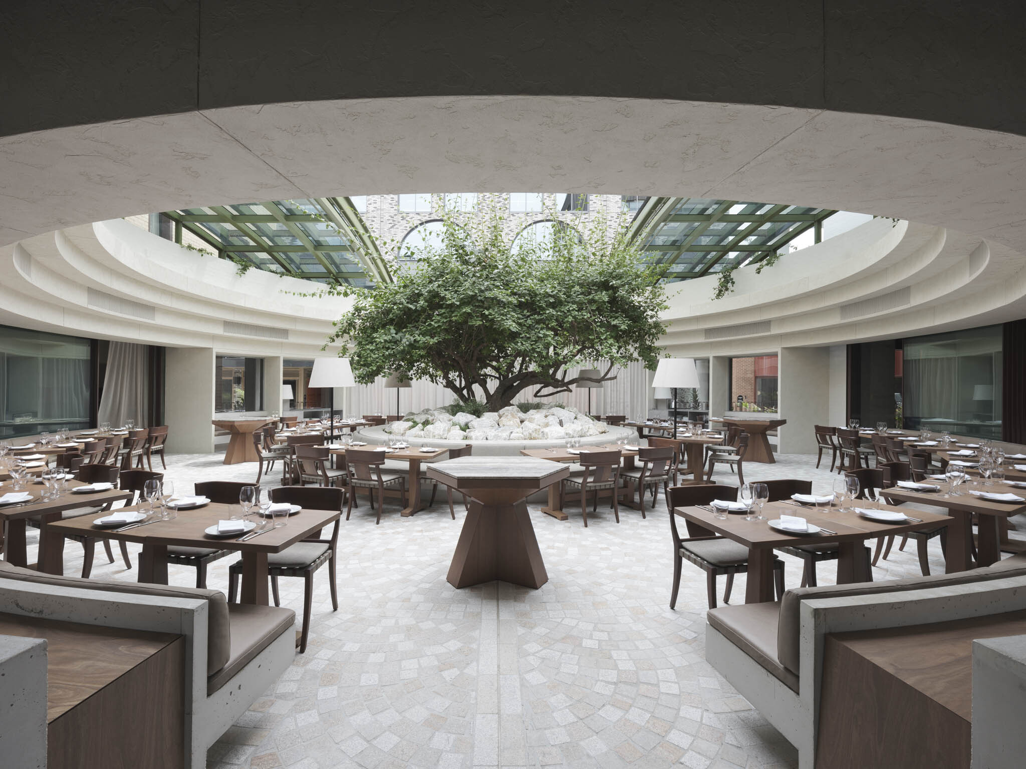 Upscale restaurant interior featuring multiple dining tables and chairs arranged around a tree under a glass-covered courtyard.