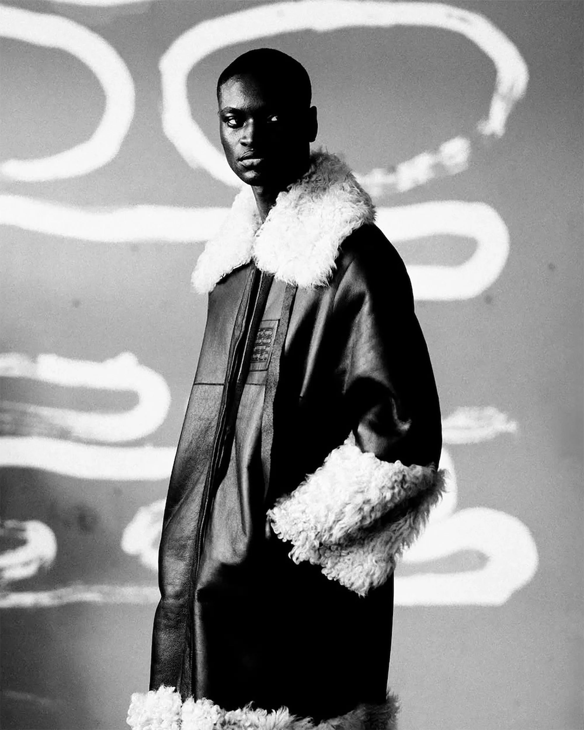 Portrait of a black male fashion model wearing a brown leather jacket with white fur trim against a grey background with white lines.