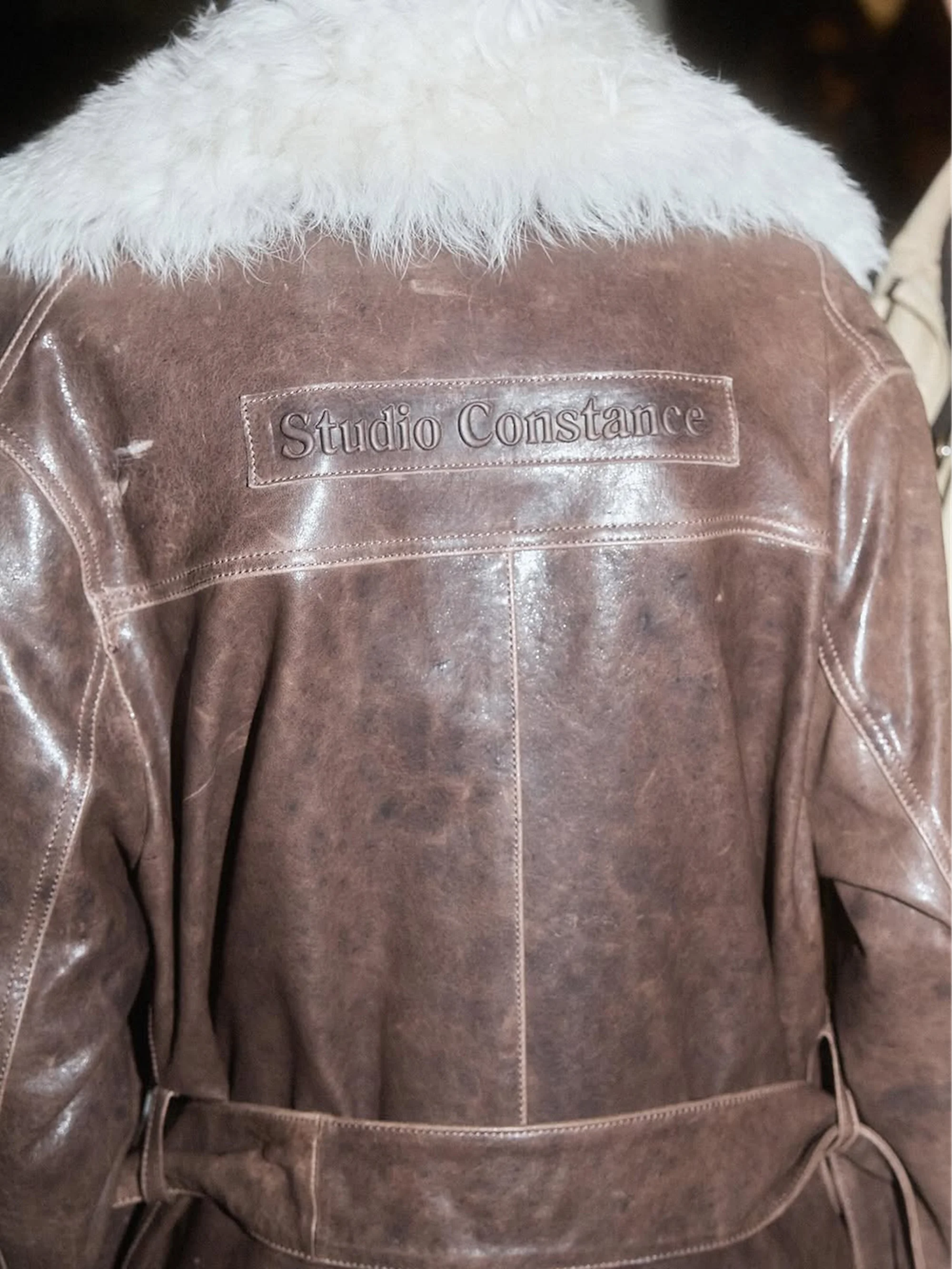 Close-up of a brown leather jacket featuring a white fur collar and 'Studio Constance' embossed on the upper back.