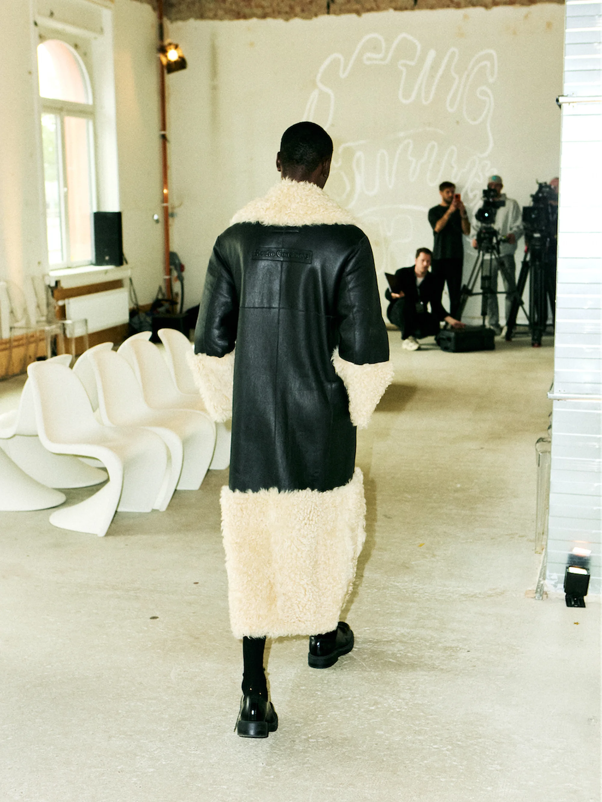 Runway fashion show featuring an adult male model in a black and cream shearling coat and black boots walking on a concrete floor.