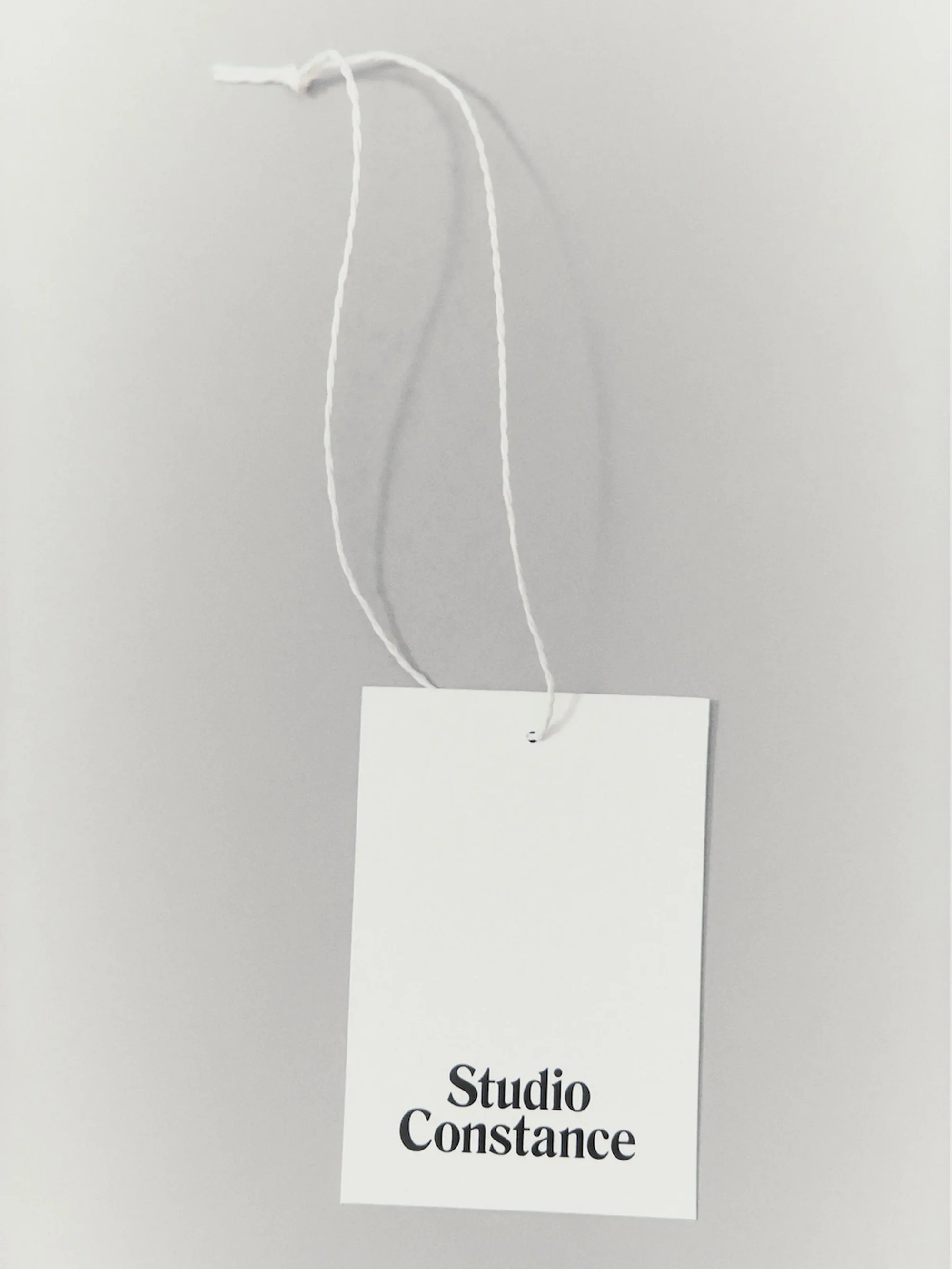 Studio Constance branded white tag hanging from a white string against a grey background in a close-up shot. Minimalist design.