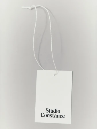 Studio Constance branded white tag hanging from a white string against a grey background in a close-up shot. Minimalist design.