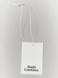 White paper hangtag with thin white string hanging against a solid grey studio background featuring the Studio Constance brand logo