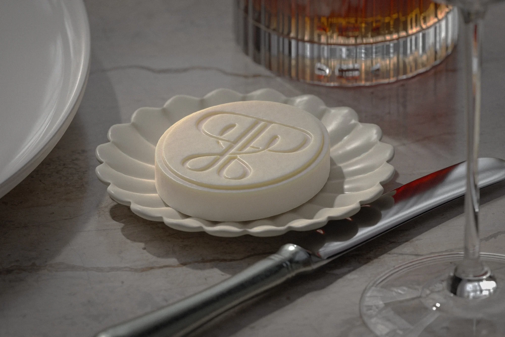 Close-up shot of a round, patterned soap on a scallop-edged dish with a butter knife and drinking glass in the background.