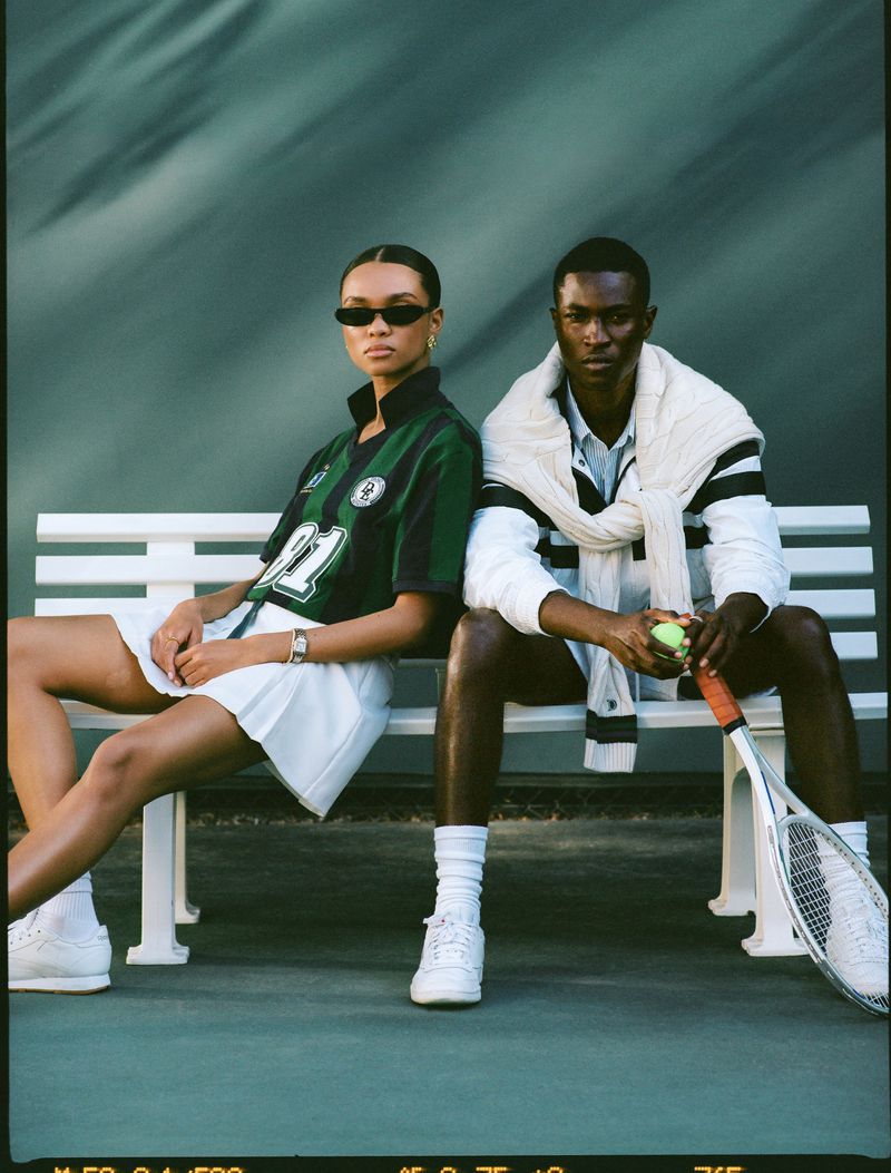 Two tennis players, a man and a woman, sit on a bench on a tennis court, the man holds a tennis ball, and the woman wears sunglasses.