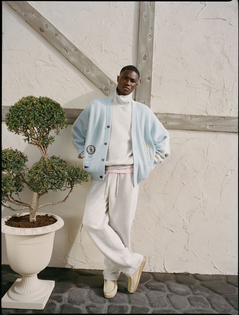 A Black male model leans against a textured wall in a light blue cardigan and white sweatpants, showcasing casual men's fashion.