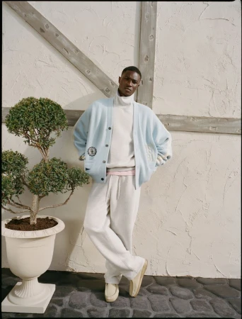 A Black male model leans against a textured wall in a light blue cardigan and white sweatpants, showcasing casual men's fashion.