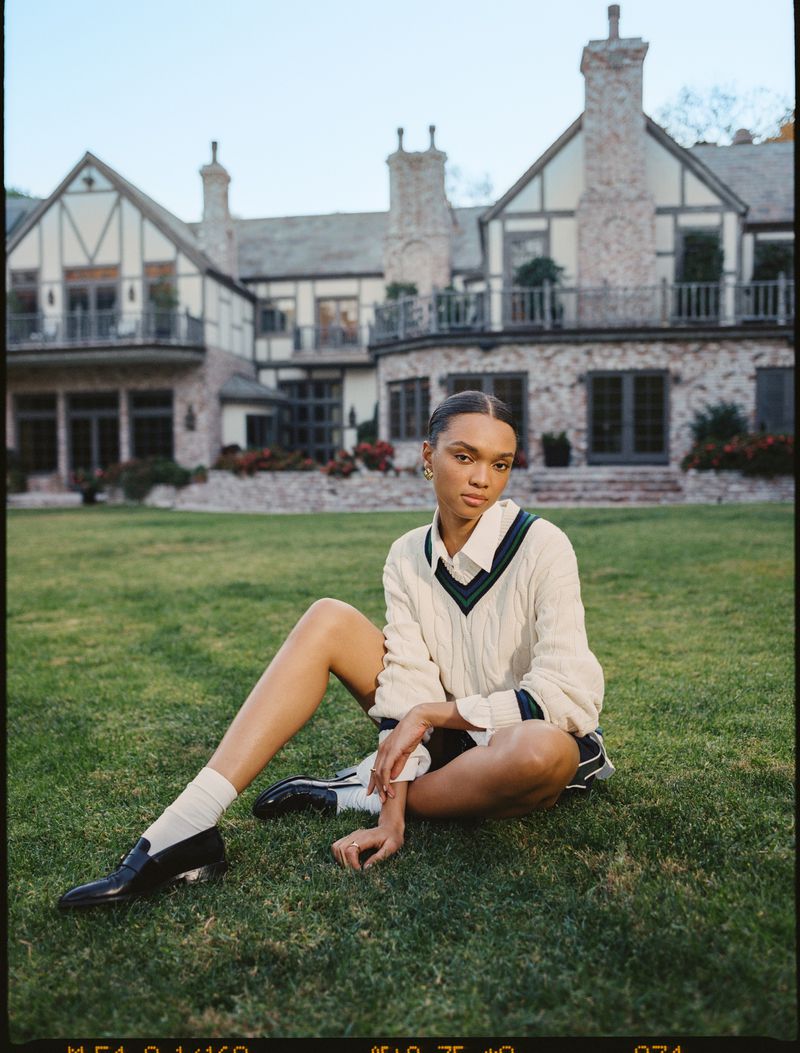 A woman poses sitting on grass in front of a large house. She wears a sweater and skirt with black loafers and white socks in a lawn setting.