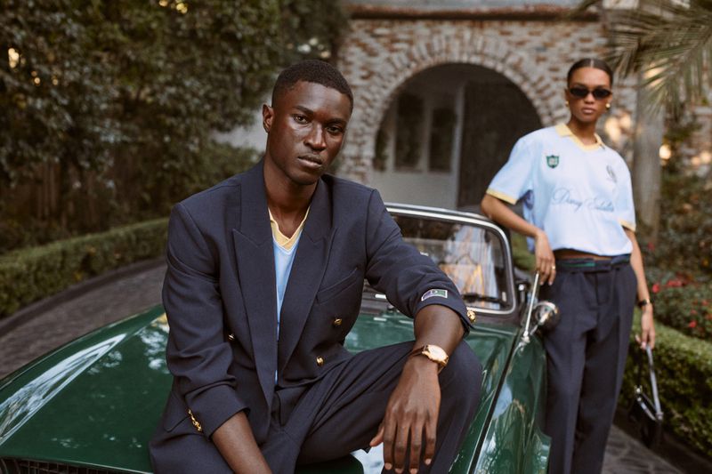 Dark-skinned male model in a navy blazer seated on a vintage green convertible with a female model standing in the background.