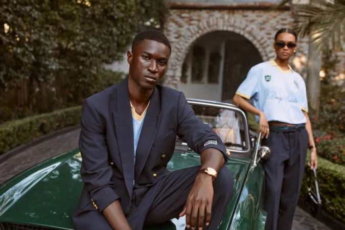 Dark-skinned male model in a navy blazer seated on a vintage green convertible with a female model standing in the background.