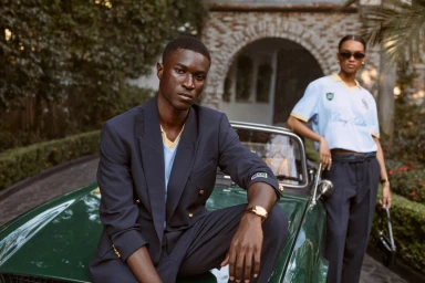 A man in a dark blazer crouches on a green vintage car while a woman stands nearby in a light blue polo shirt.