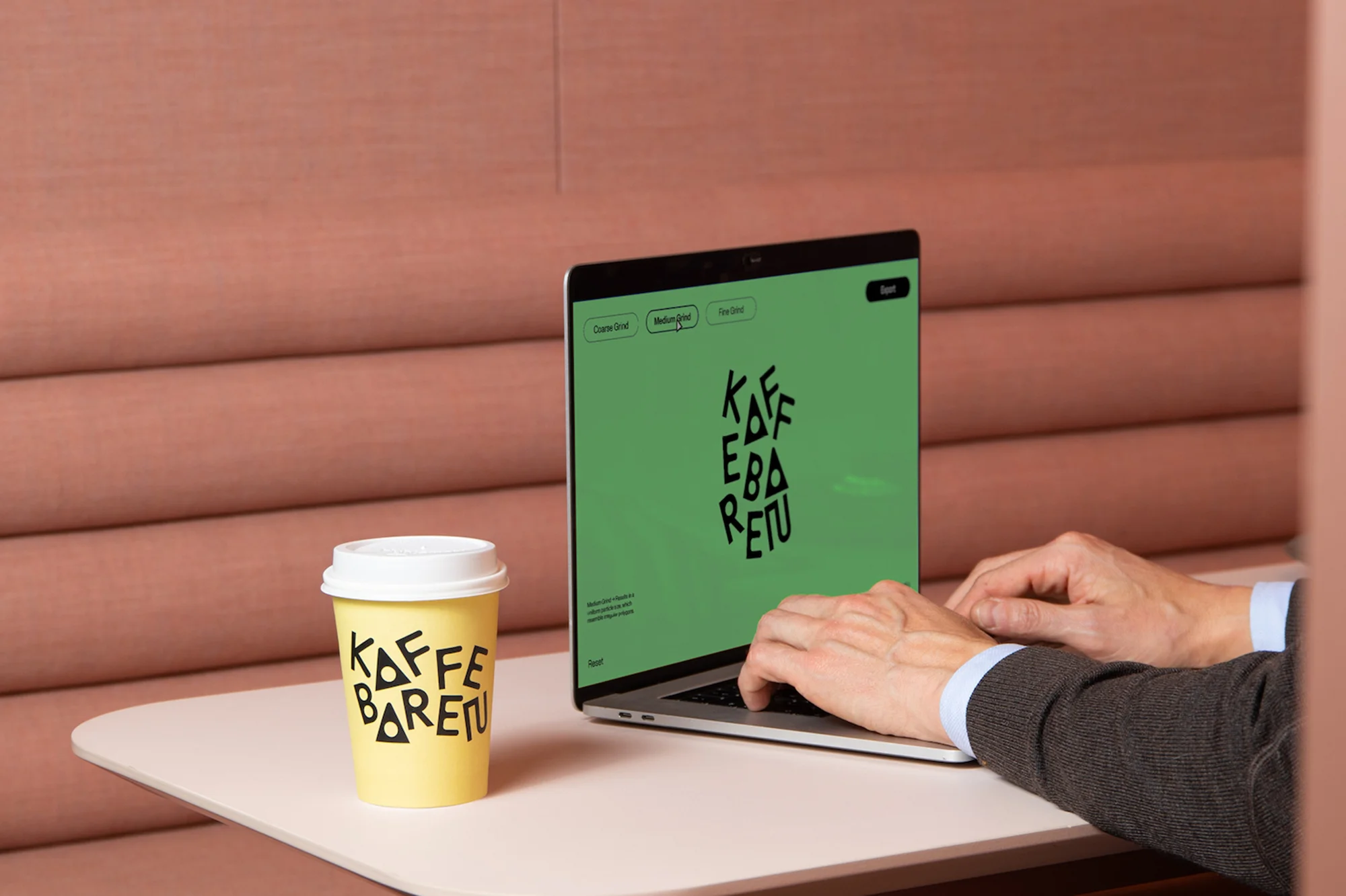 Person works at a laptop with coffee cup on a table in a modern office setting, displaying Kaffe Baretu logo on the screen and cup.