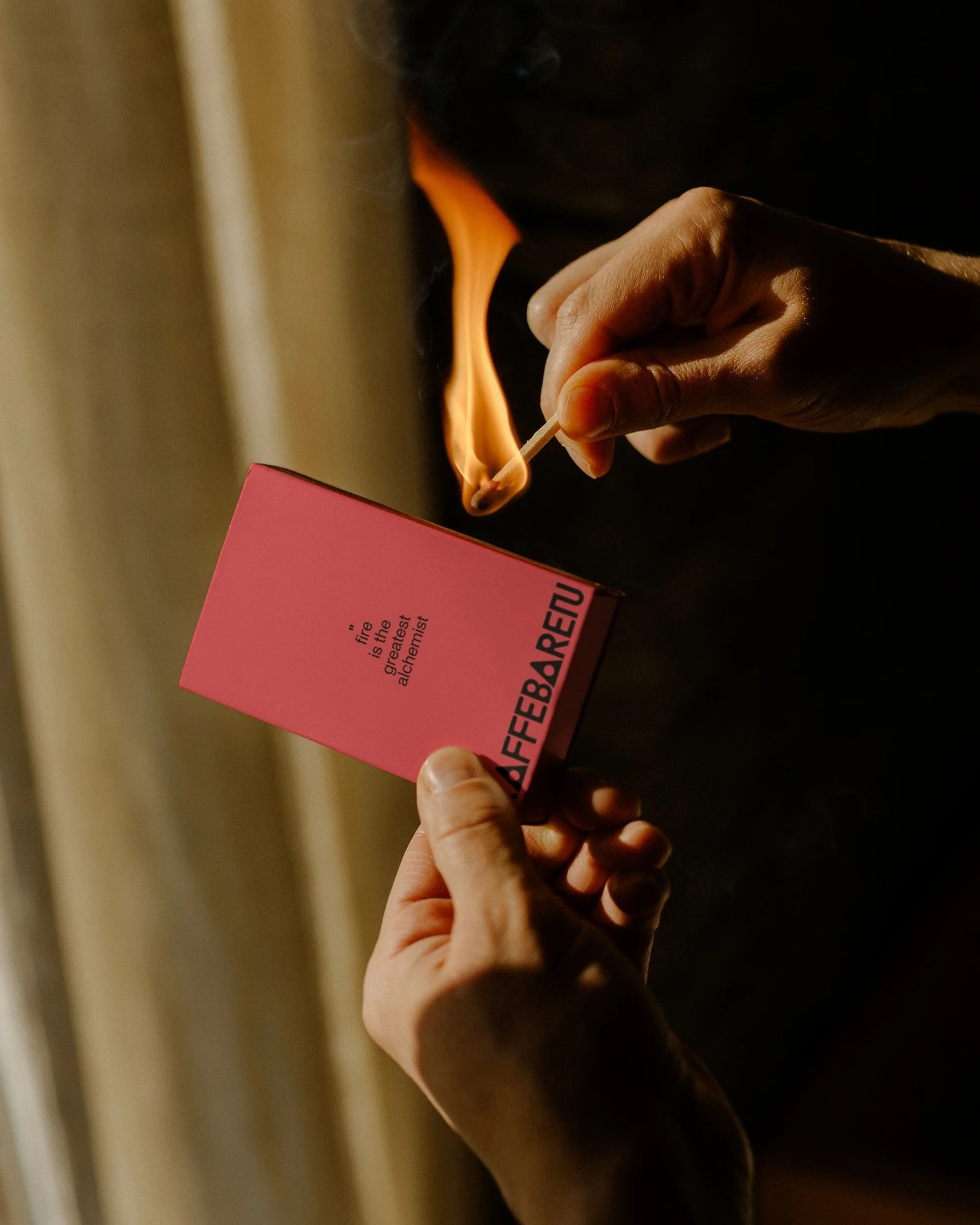 Hands strike a match against a matchbox in a dimly lit space with beige curtain; creating a fiery ambiance.