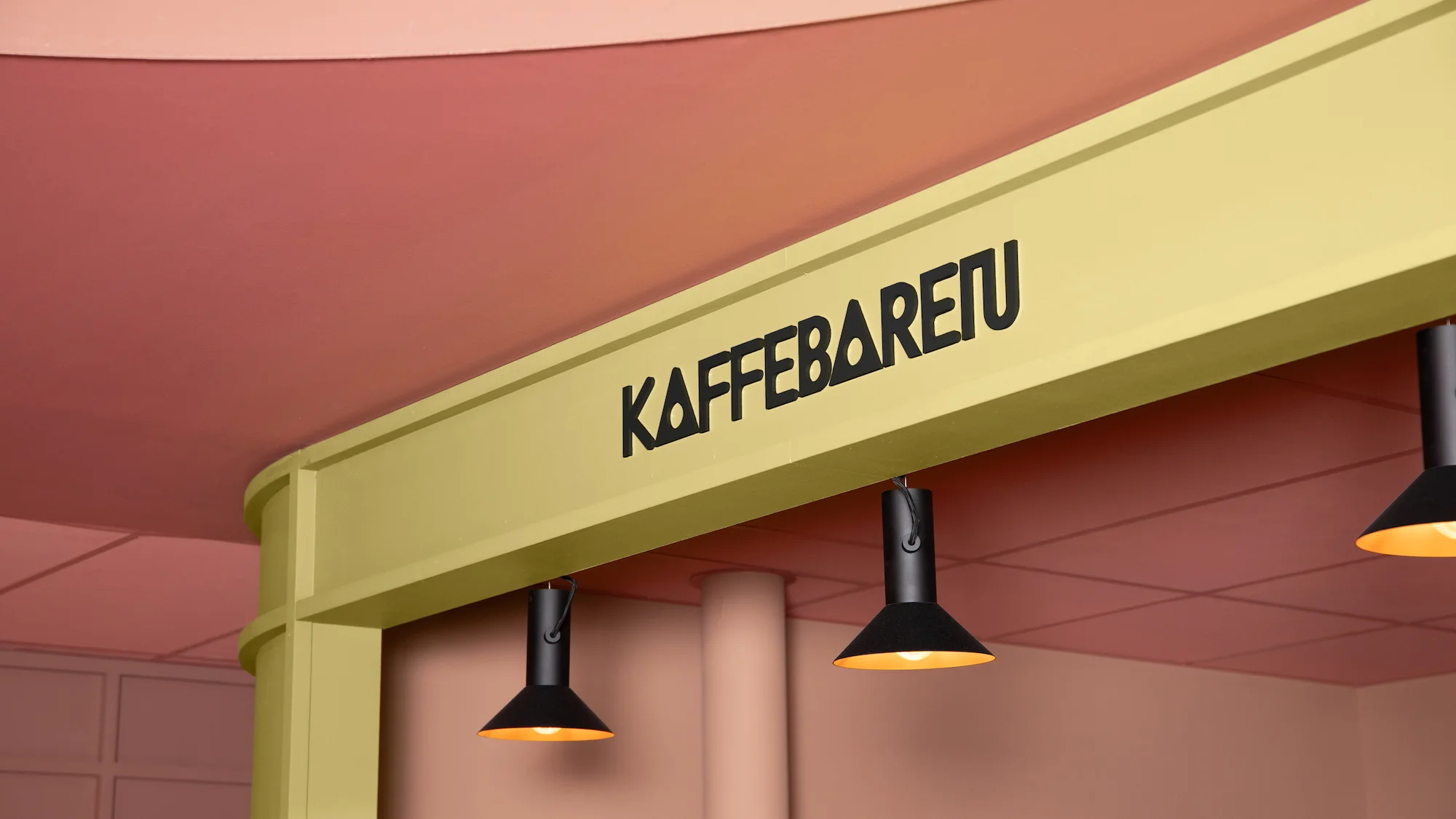 Interior view of a cafe with black pendant lights and the KaffeBaretU sign above a light yellow frame against a pink ceiling.