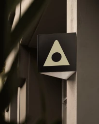 Square black sign with a light triangle and dark circle logo mounted on a pale wall outdoors.