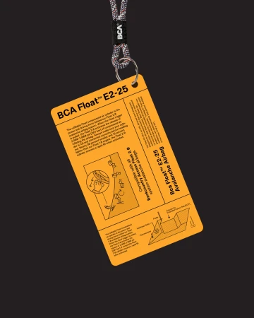 Yellow BCA Float E2-25 Avalanche Airbag instructional card with diagrams hangs from a lanyard against a dark background. 