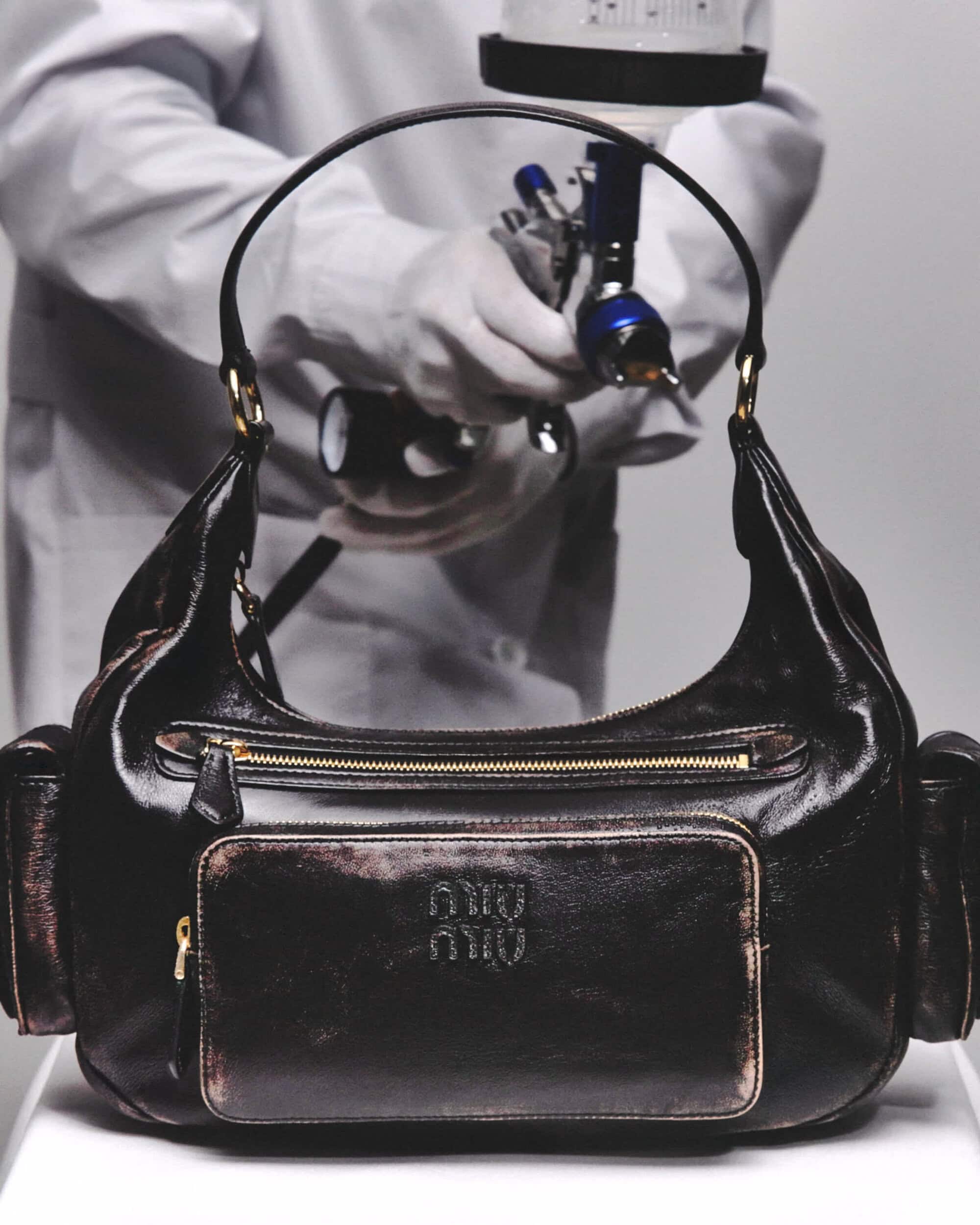 Close-up of a black leather Miu Miu handbag, being spray-painted by a person in a white lab coat and gloves in a photo studio.