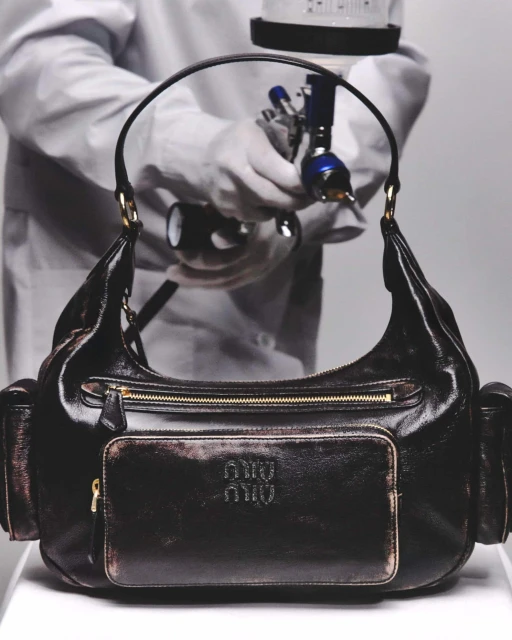 Close-up of a black leather Miu Miu handbag, being spray-painted by a person in a white lab coat and gloves in a photo studio.