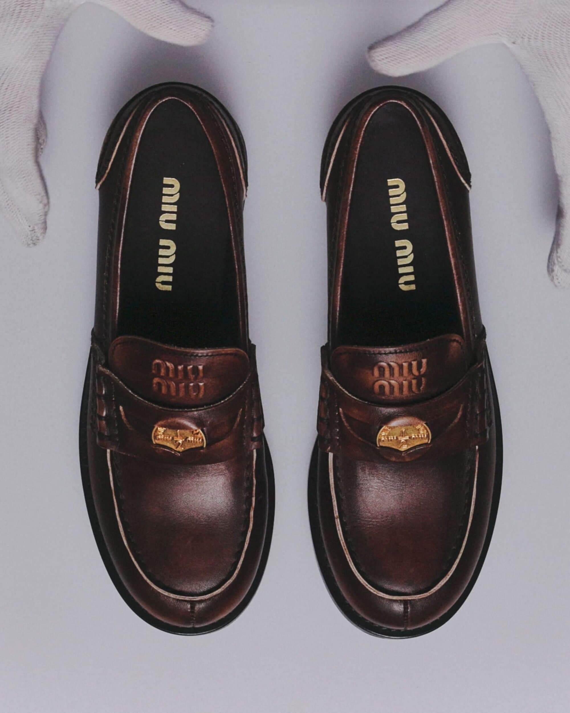 Miu Miu brown leather loafers featuring a gold-toned emblem on a grey surface, held in place by hands in white gloves.