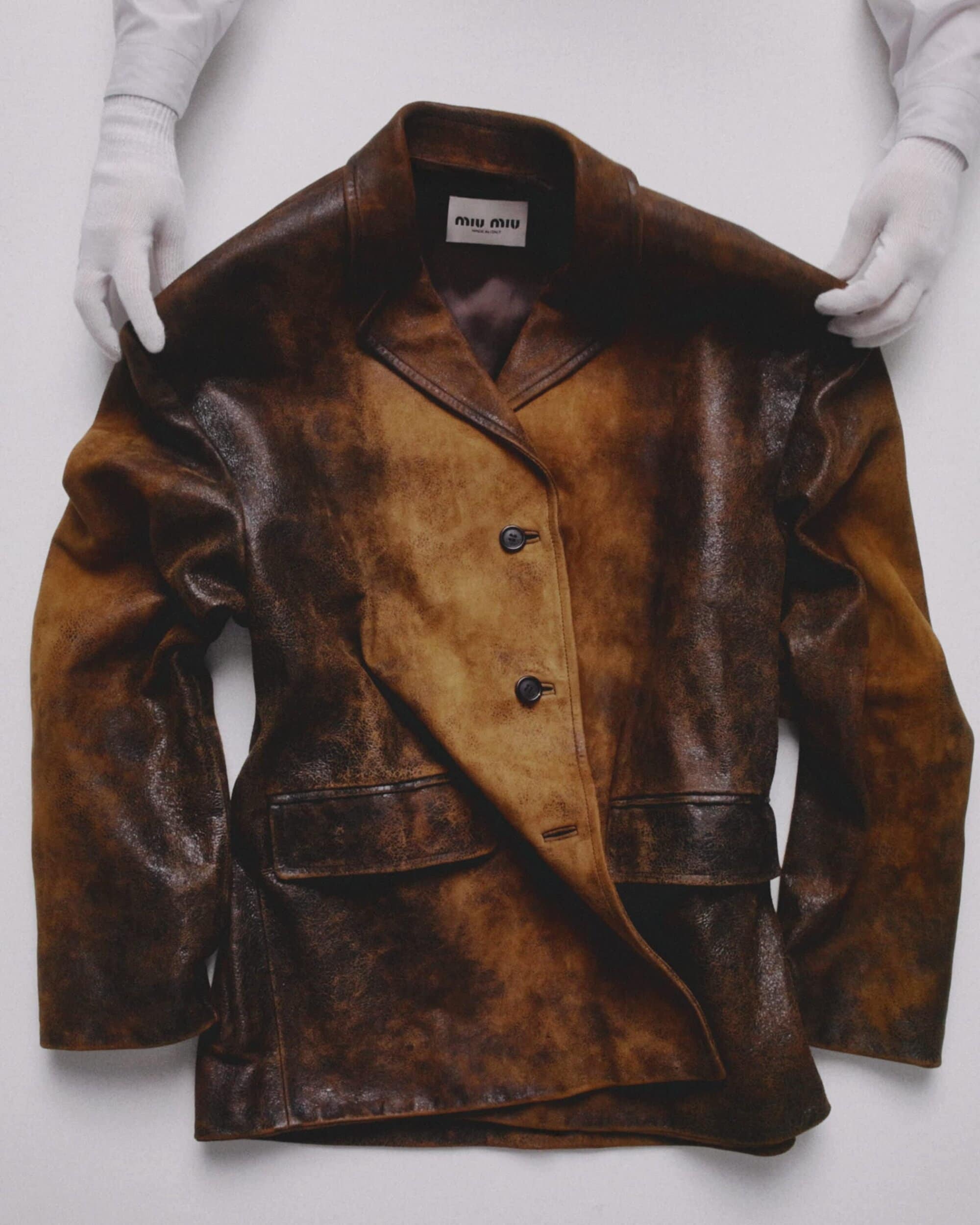 Close-up of a distressed brown leather jacket with gloved hands holding it open, brand logo 'Miu Miu' is visible on the inner label.
