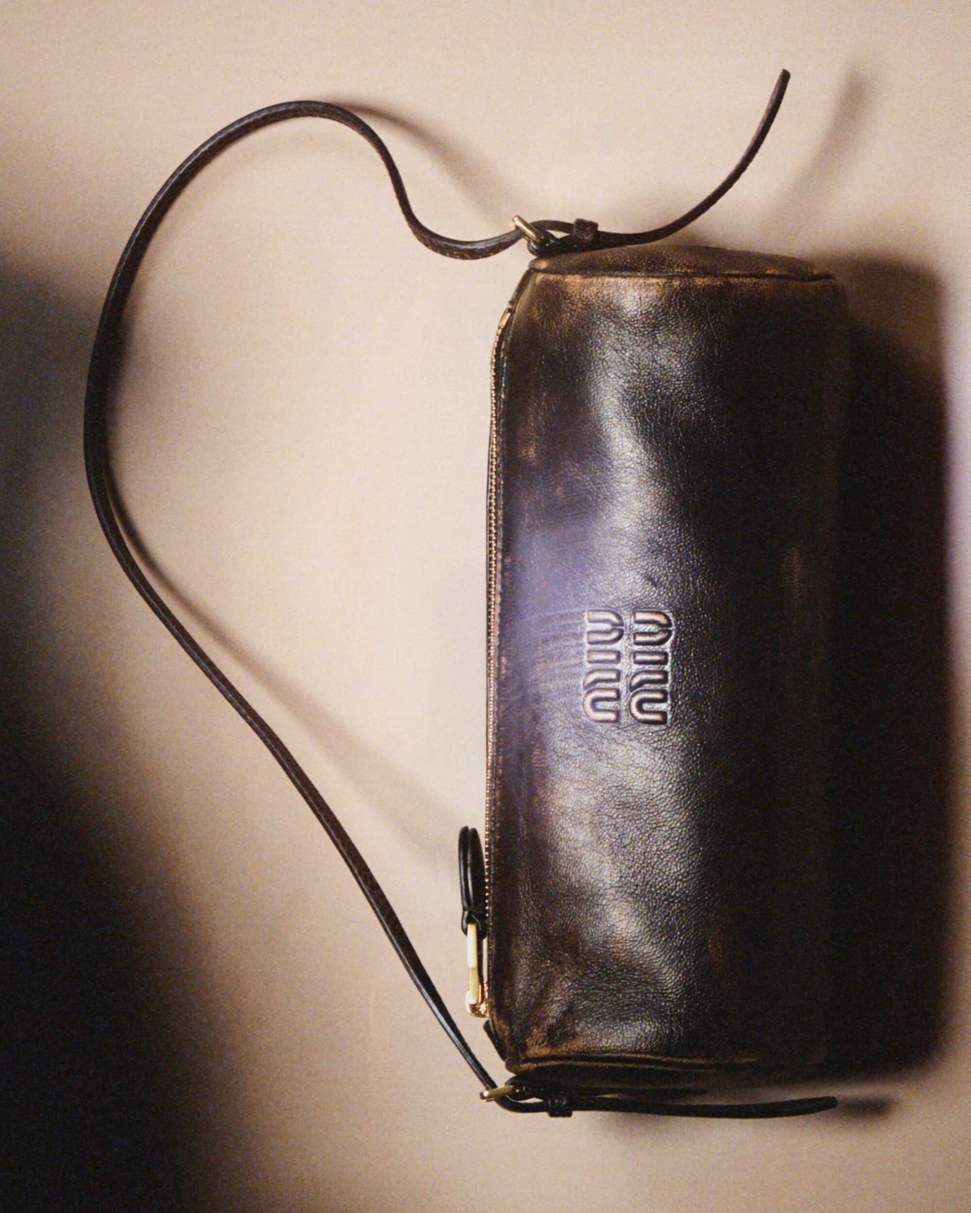Brown leather cylindrical bag with shoulder strap and metal Miu Miu logo detail against a neutral beige background.