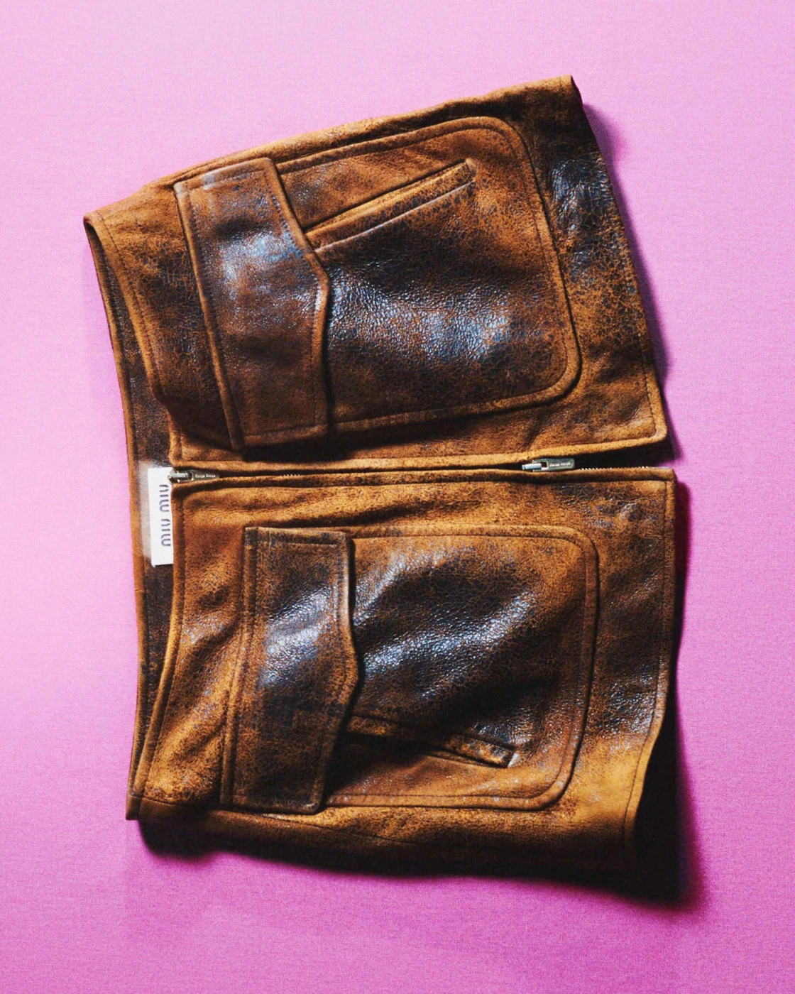 Distressed brown leather jacket garment laid flat against a bright pink background for Miu Miu ad campaign