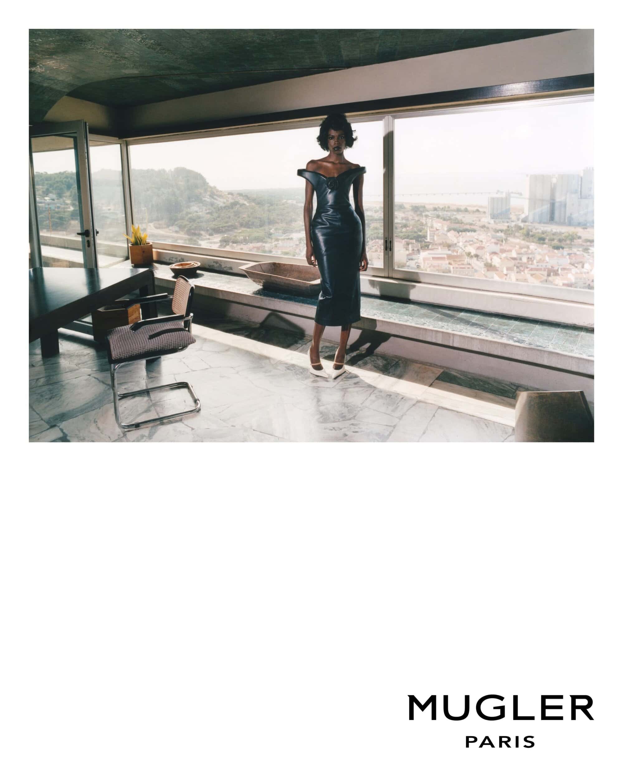 An adult woman wearing a black dress poses inside a modern home with large windows overlooking a cityscape and distant mountains.
