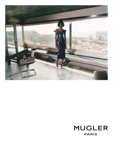 An adult woman wearing a black dress poses inside a modern home with large windows overlooking a cityscape and distant mountains.