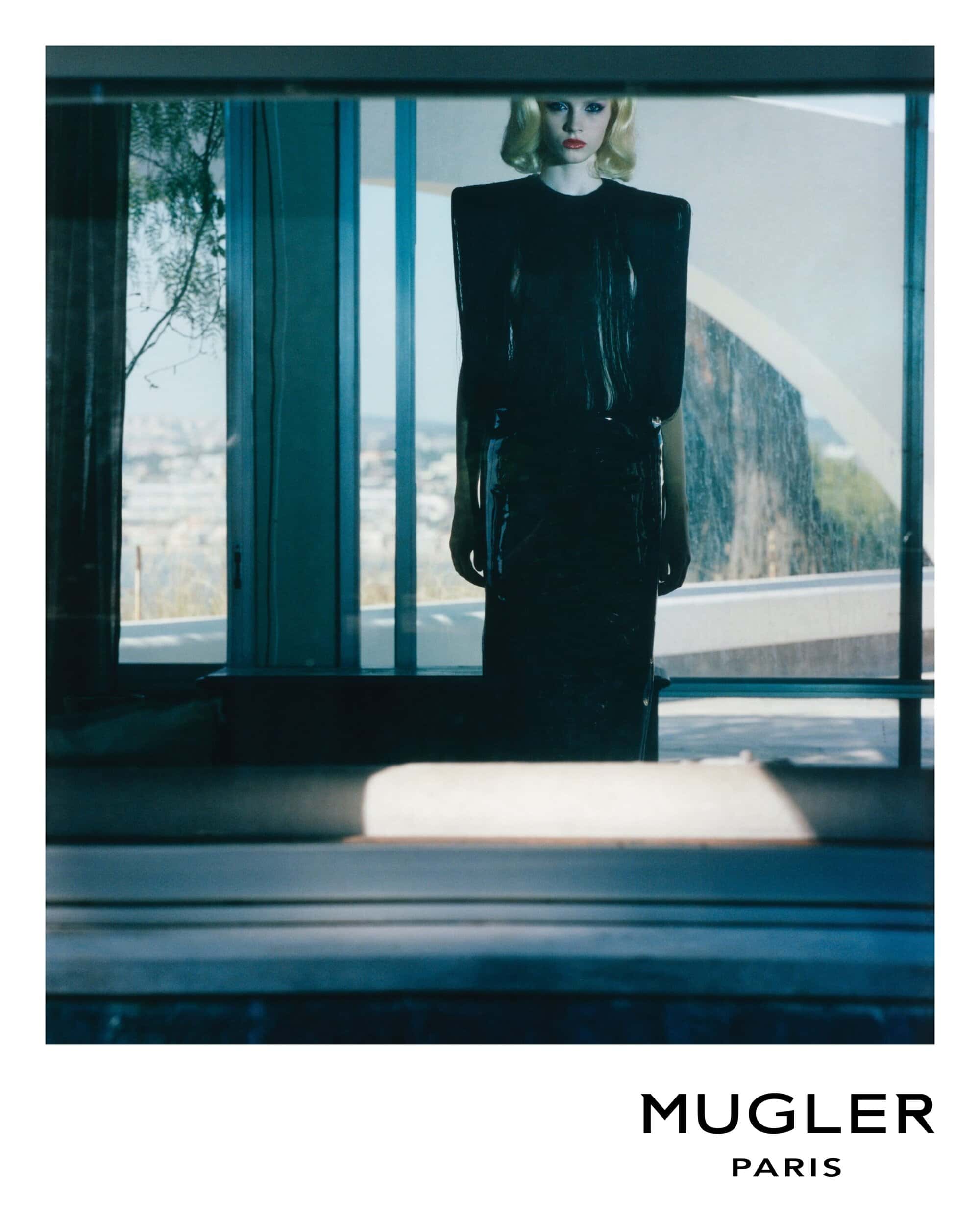 Blonde fashion model in glossy black dress with padded shoulders in a bright, modern home setting for a Mugler fashion advertisement.