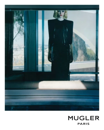 Blonde fashion model in glossy black dress with padded shoulders in a bright, modern home setting for a Mugler fashion advertisement.