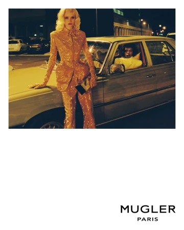 Fashion shot of a woman in a sequined suit leaning on a car, another person sits inside the car in an urban environment at night.