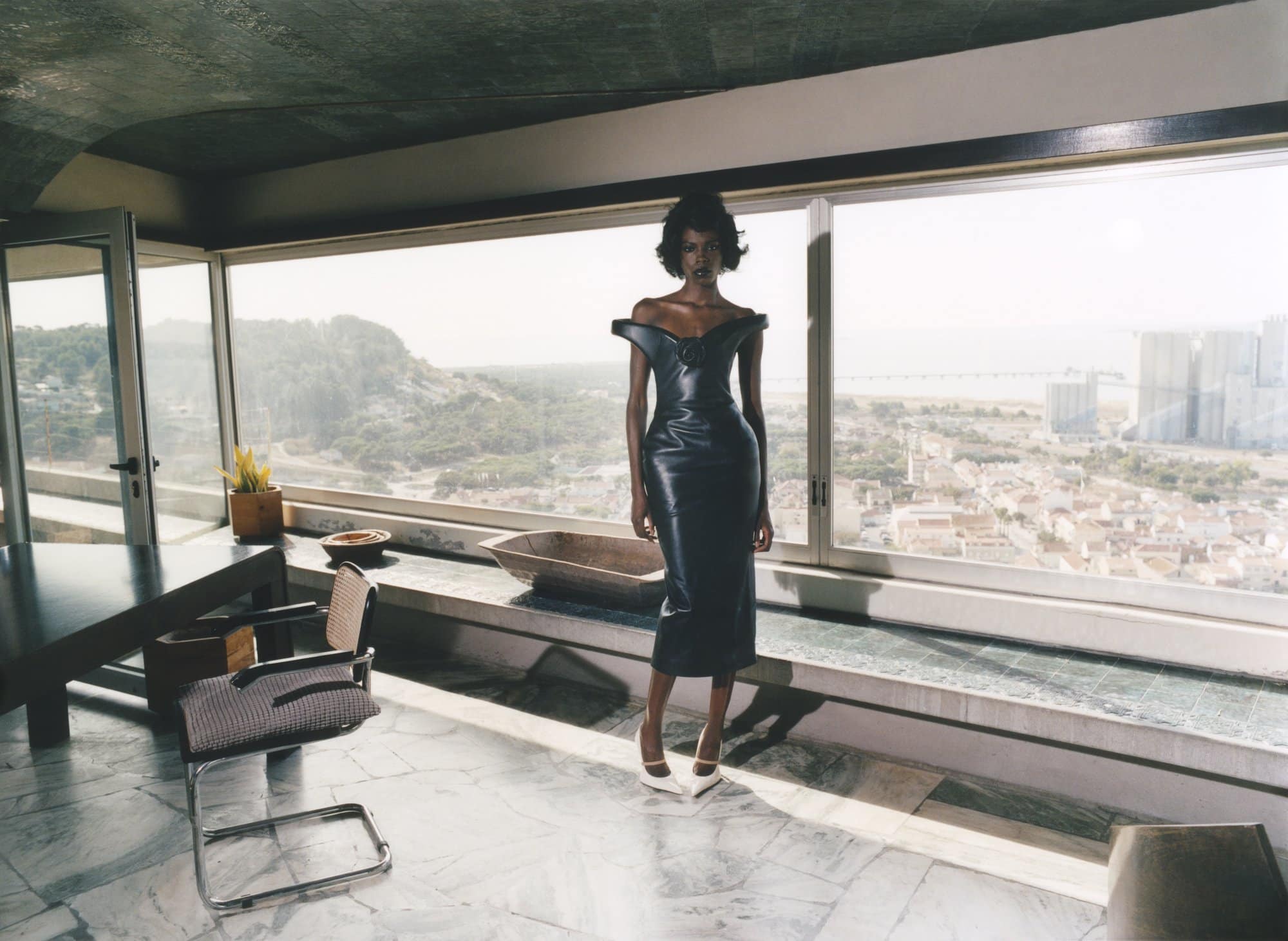 Elegant dark-skinned woman in black dress and white heels stands in an upscale apartment with a city and mountain view.