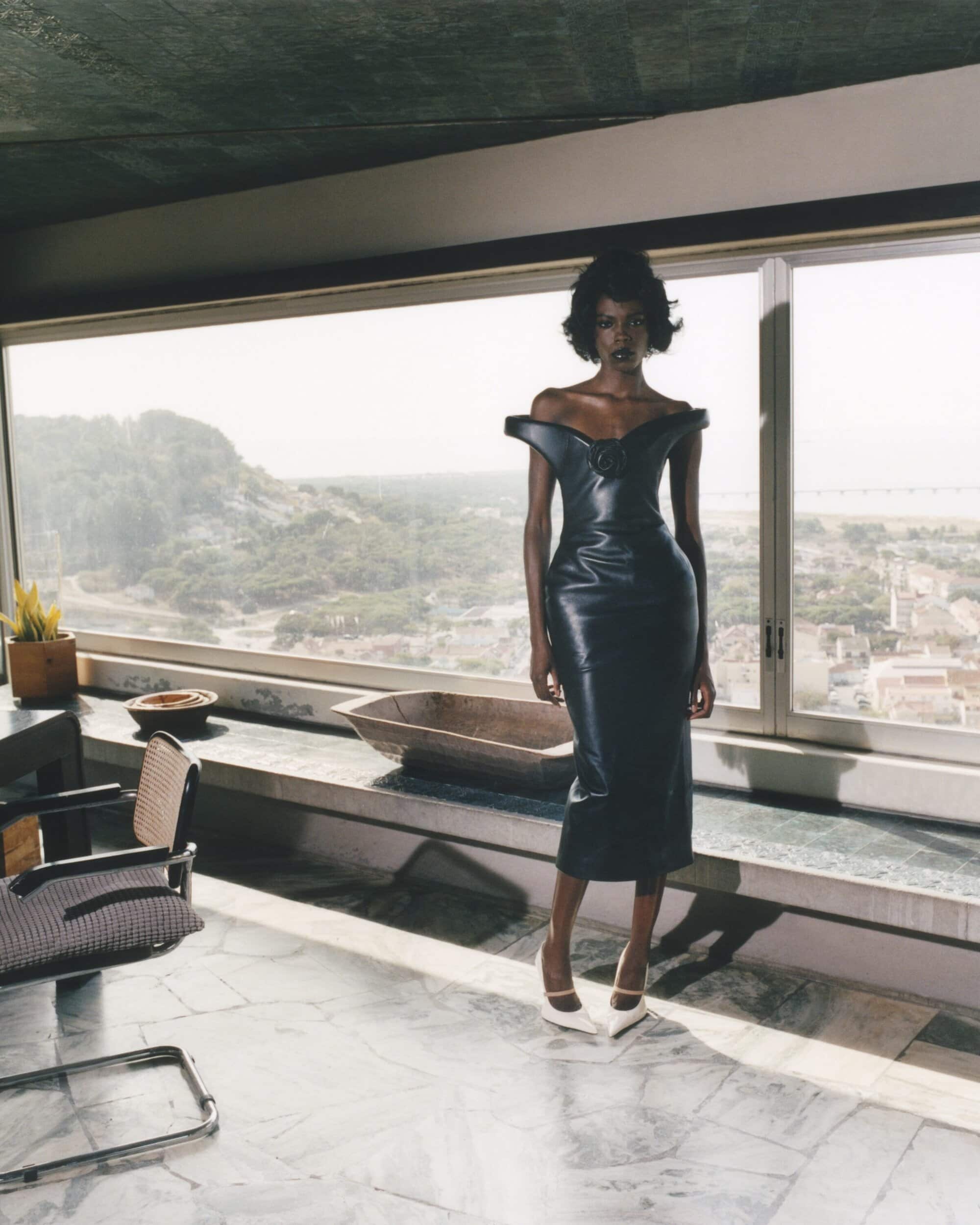 Indoor studio portrait of an adult woman posing in a fitted black dress and white heels against a scenic window backdrop.