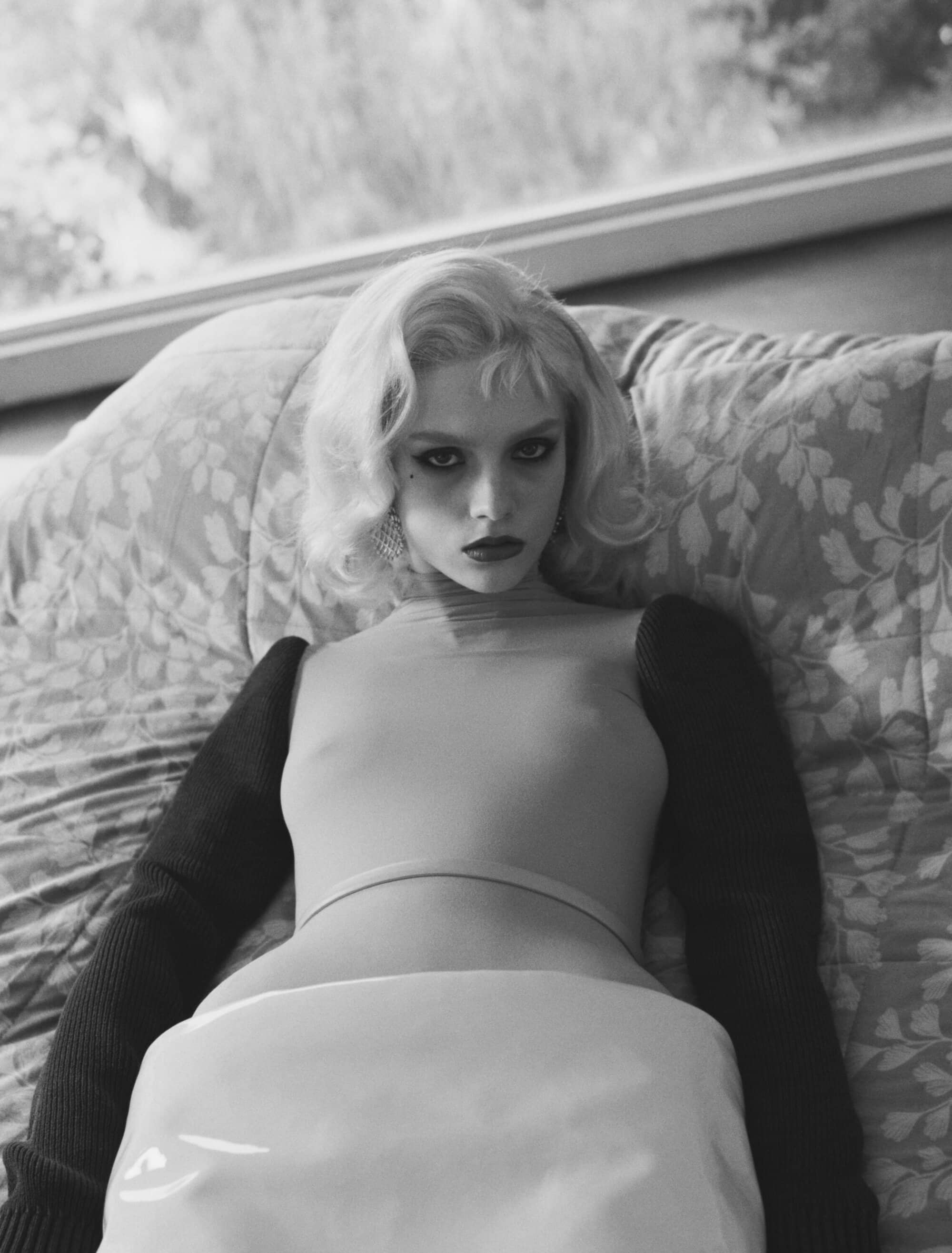 Monochrome portrait of a woman with blonde wavy hair lying on a patterned surface wearing a top and arm sleeves, and a garment.