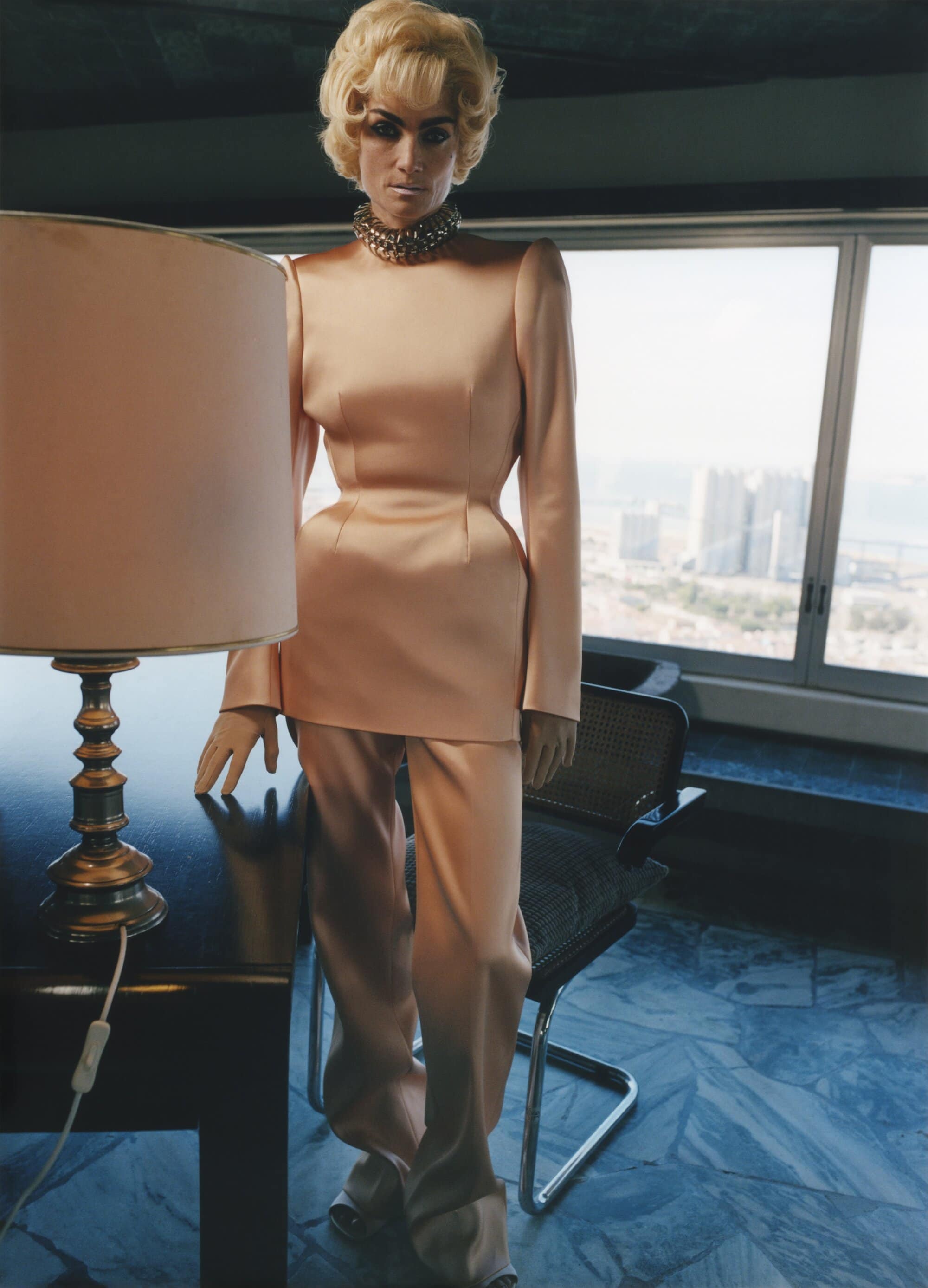 An adult woman with blonde hair is styled in a pale pink pantsuit, complemented by a jeweled necklace in an indoor setting near windows.