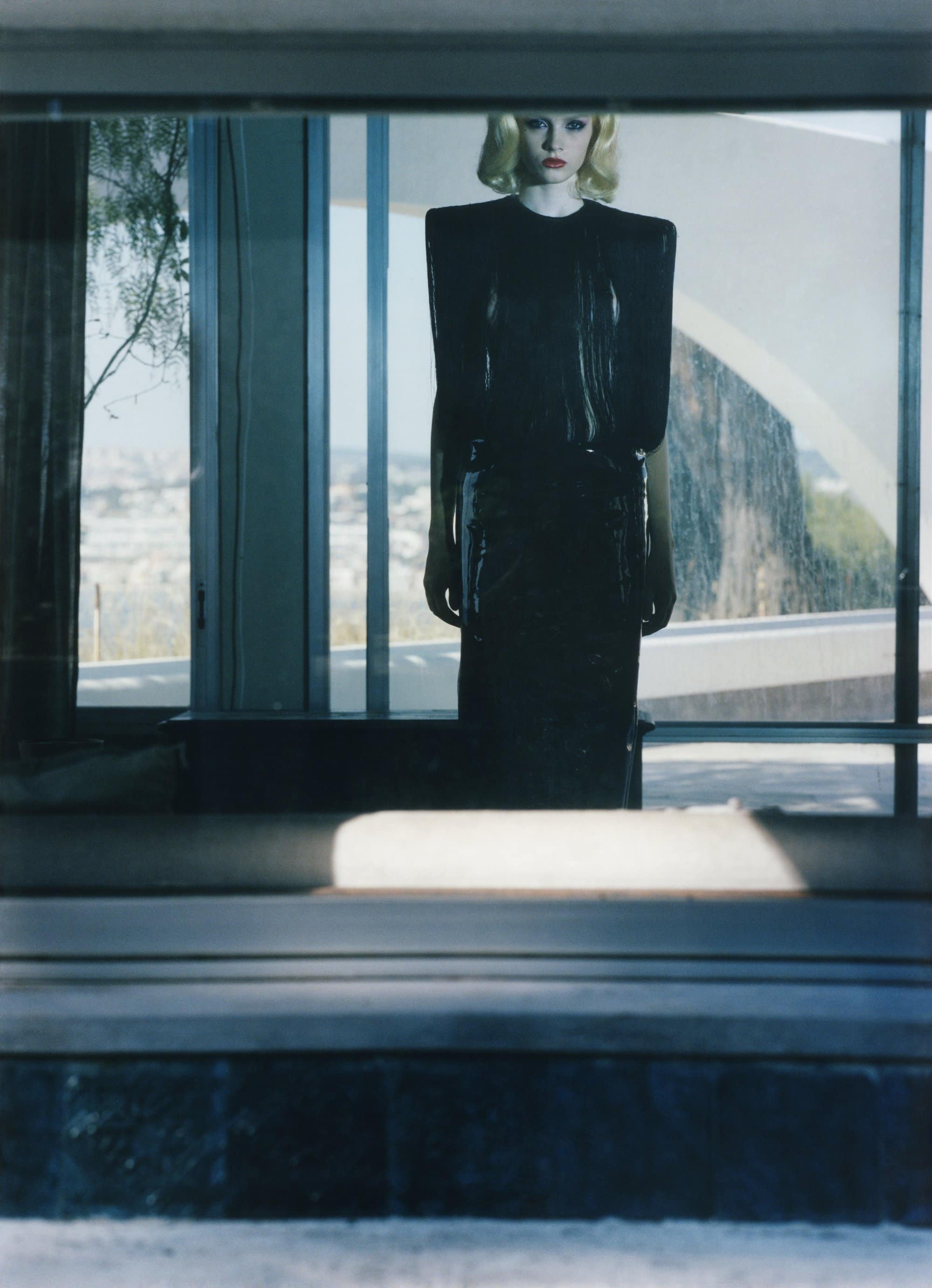 Blonde woman in a black dress stands indoors, framed by a window with a bright outdoor view, creating a reflective and modern aesthetic.