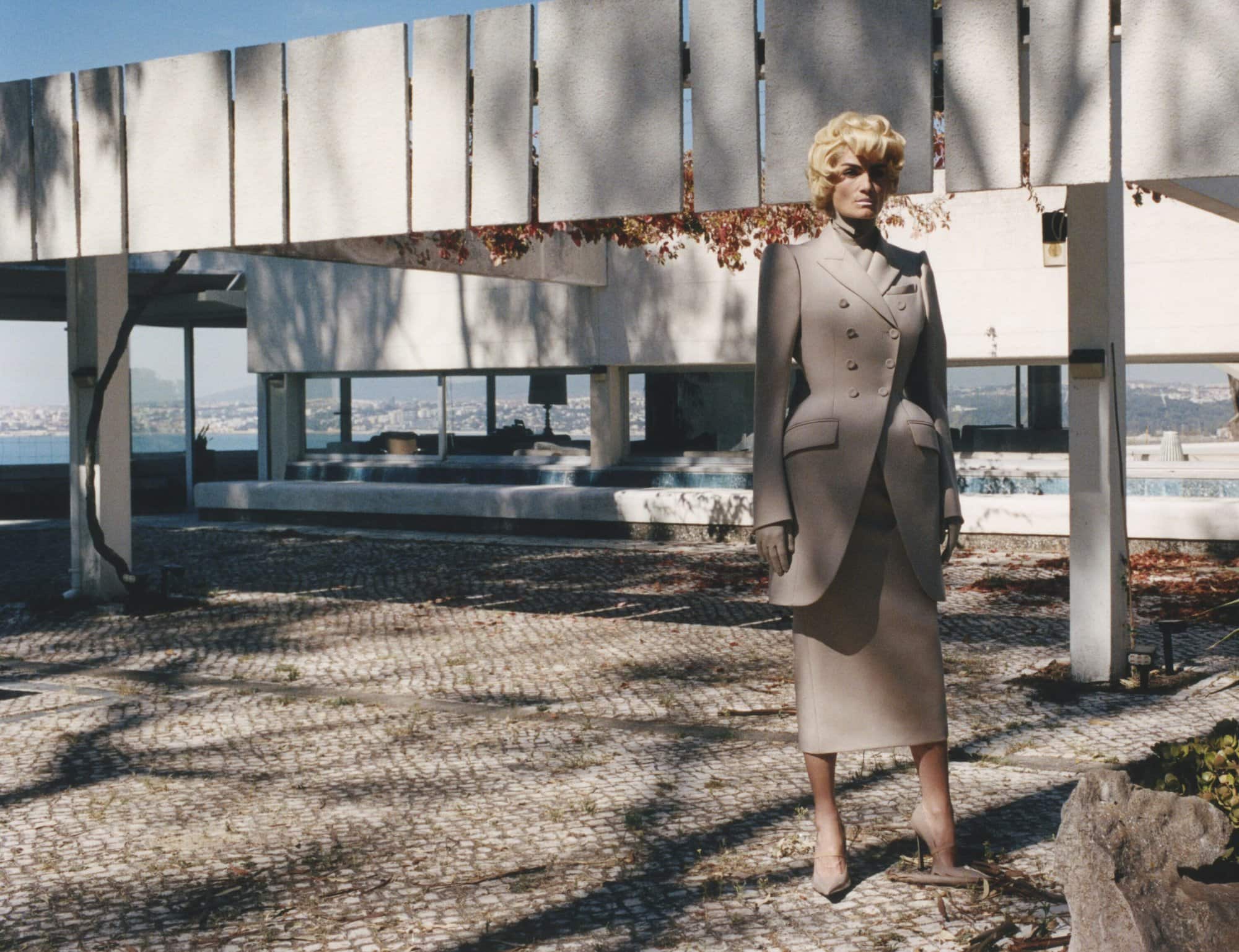 An adult female wearing a light grey suit and high heels stands in a courtyard near a modern white building with large windows.