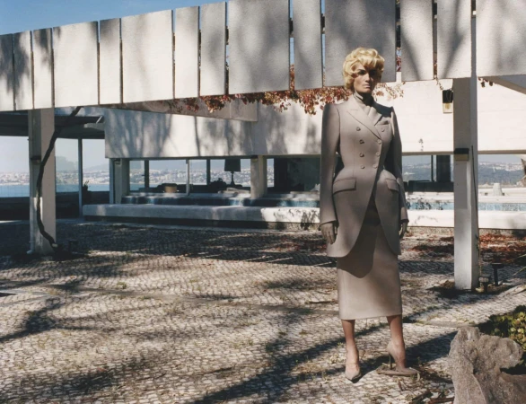 An adult female wearing a light grey suit and high heels stands in a courtyard near a modern white building with large windows.