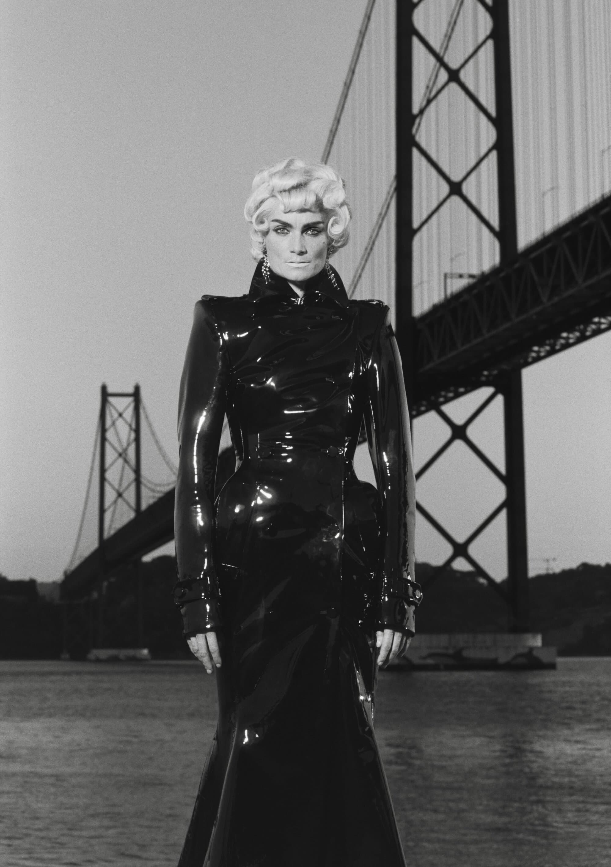 Monochrome shot of a fair-skinned adult female fashion model wearing a shiny black trench coat with a bridge in the background.