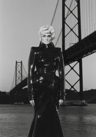 Monochrome shot of a fair-skinned adult female fashion model wearing a shiny black trench coat with a bridge in the background.